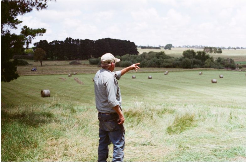 An image of a farmer standing in a field, pointing towards the horizon. The field is dotted with round hay bales under a partly cloudy sky